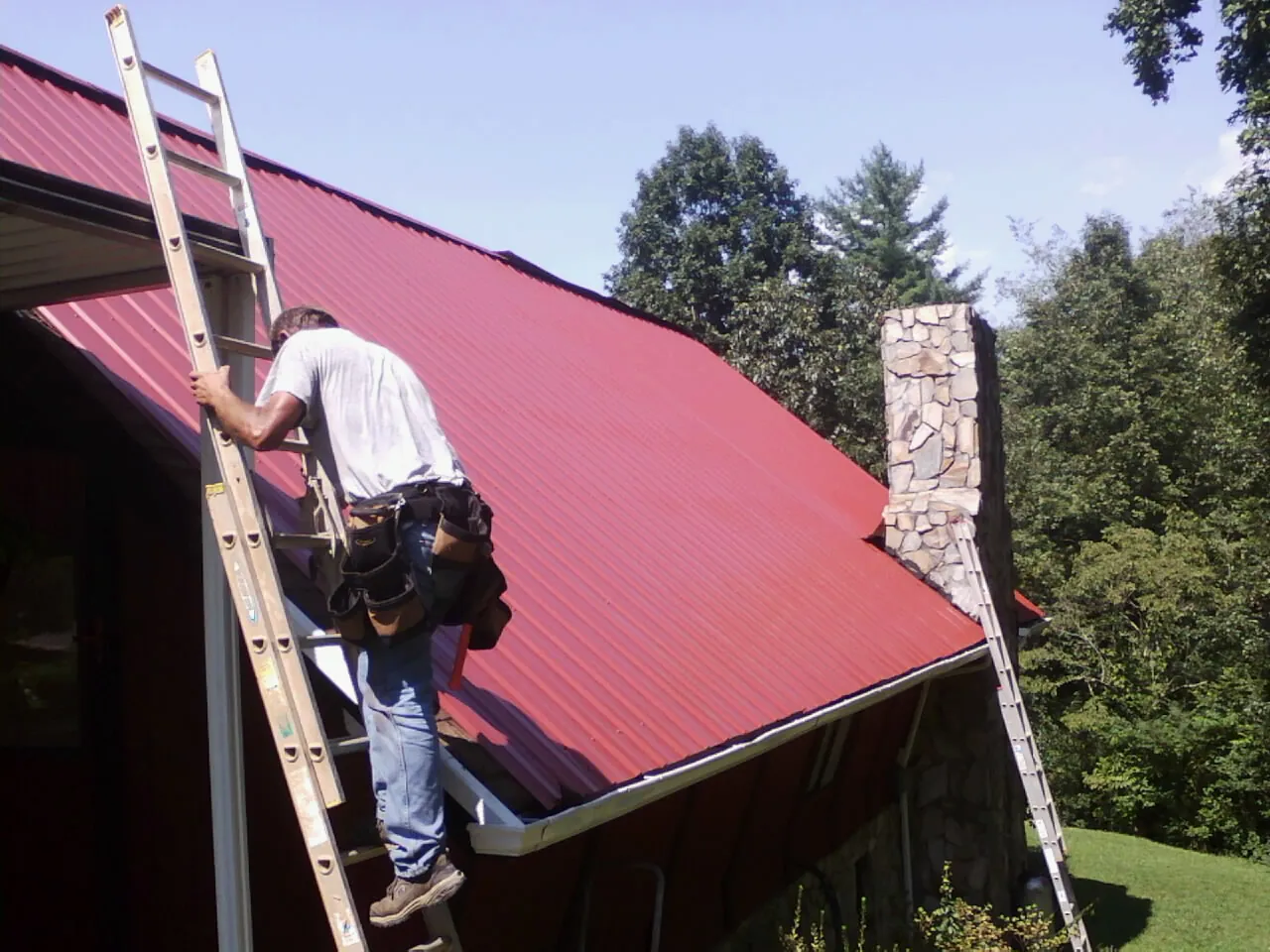 Roofer on a ladder beside a red metal roof and stone chimney — Soffit & Fascia Repair by Roofing Brothers in Connell